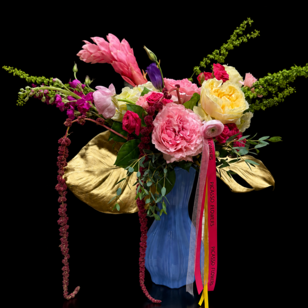Designer Mother’s Day flower arrangement in a blue vase with pink peonies, yellow roses, hot pink roses, white hydrangeas, gold tropical leaves, cascading amaranthus, eucalyptus, and Picasso Flowers ribbon.