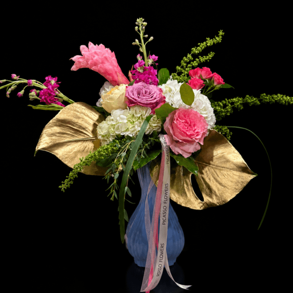 Elegant Mother’s Day flower arrangement in a blue vase featuring pink roses, white hydrangeas, magenta flowers, green accents, gold tropical leaves, and Picasso Flowers ribbon